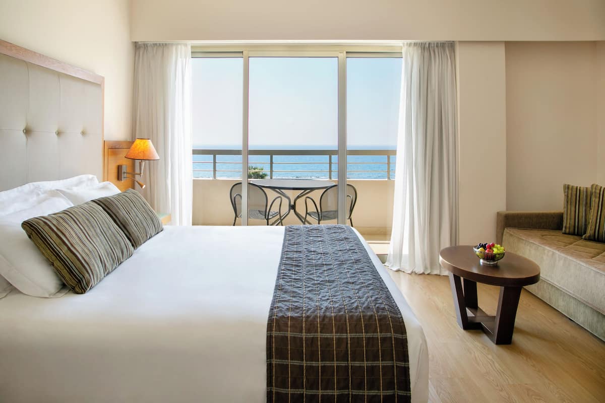 Quarto de hotel com uma cama grande, varanda com mesa e cadeiras, e vista para o mar.