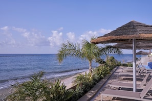 Un resort frente a la playa con una estructura de techo de paja, sillas de descanso y vista al mar.
