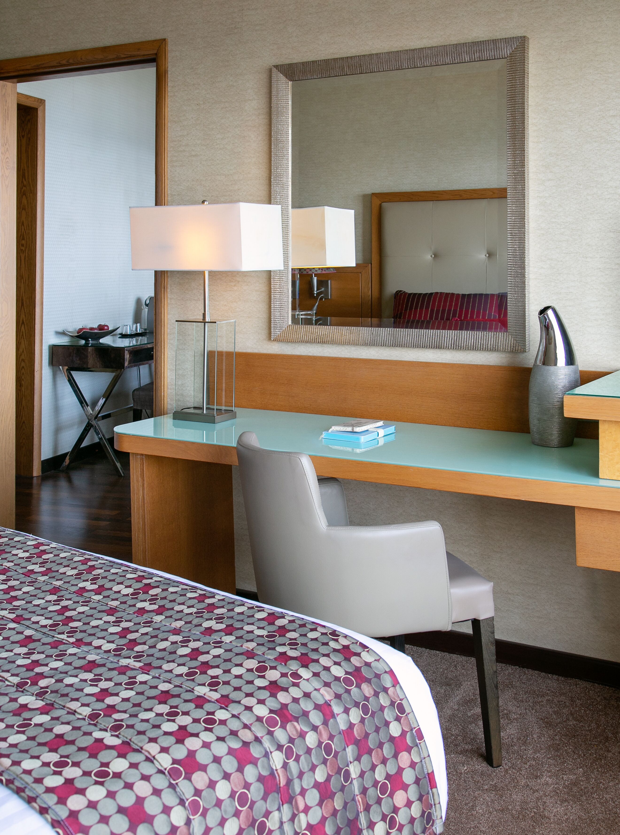 A hotel room with a bed, a desk, a chair, a lamp, a mirror, and a vase.