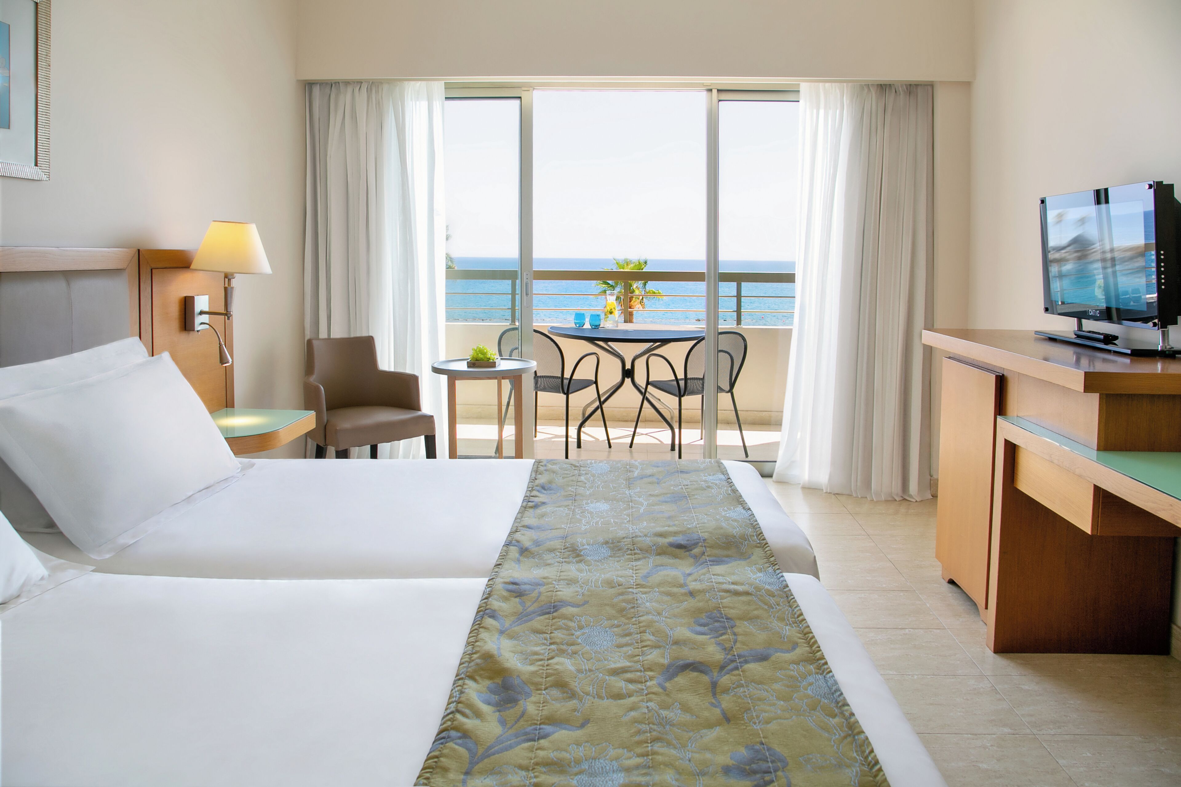 A hotel room with a bed, a desk, a television, a chair, a table with a plant, and a view of the ocean.