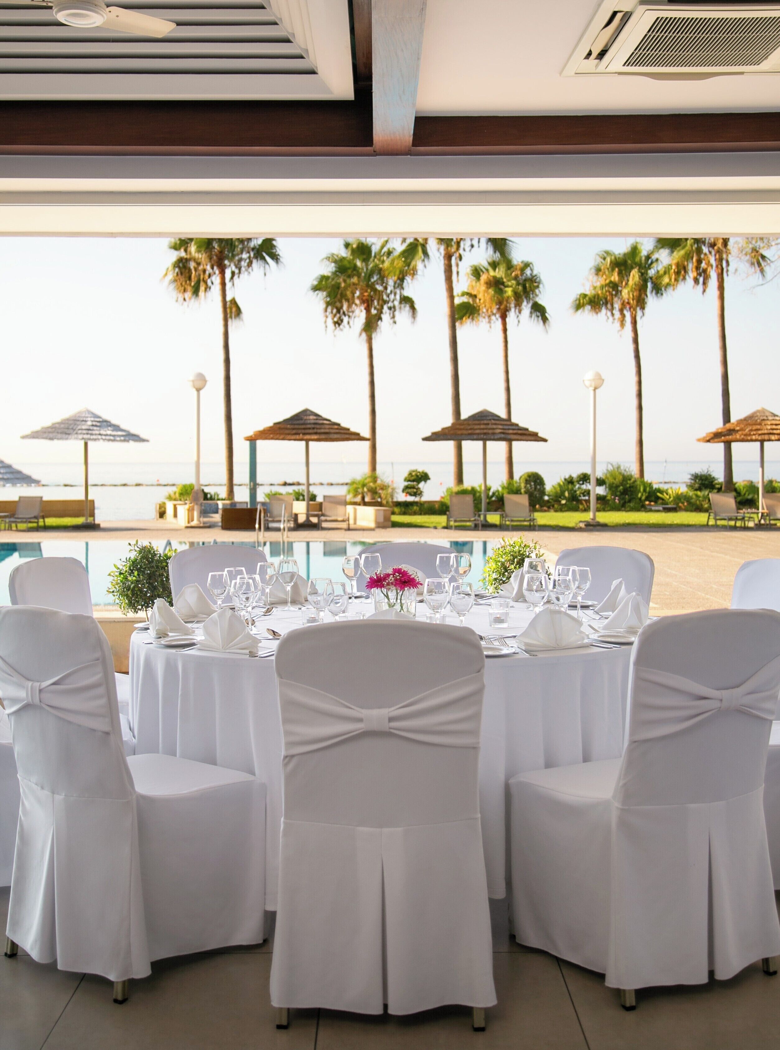 A dining area with tables set for a meal, overlooking a beach with palm trees and umbrellas.