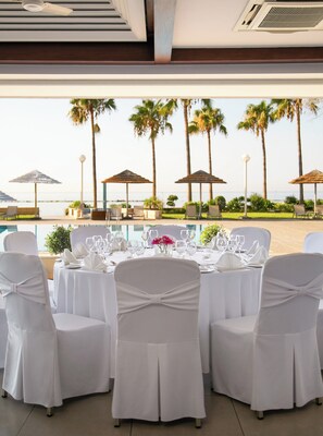 A dining area with tables set for a meal, overlooking a beach with palm trees and umbrellas. - Atlantica Miramare Beach (Limassol)