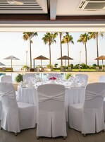 Comedor con mesas dispuestas para una comida, con vista a una playa con palmeras y sombrillas.