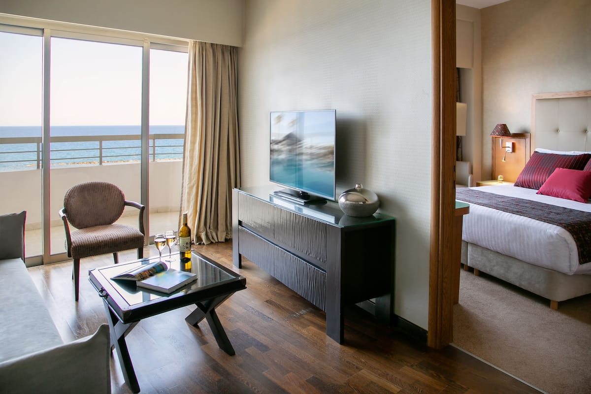 Quarto de hotel com uma janela grande, televisão, cama, mesa de cabeceira e uma cadeira.