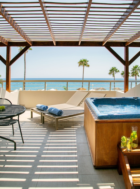 Uma área de estar coberta ao ar livre, com vista para o oceano, uma banheira de hidromassagem e uma mesa com uma cesta de frutas.