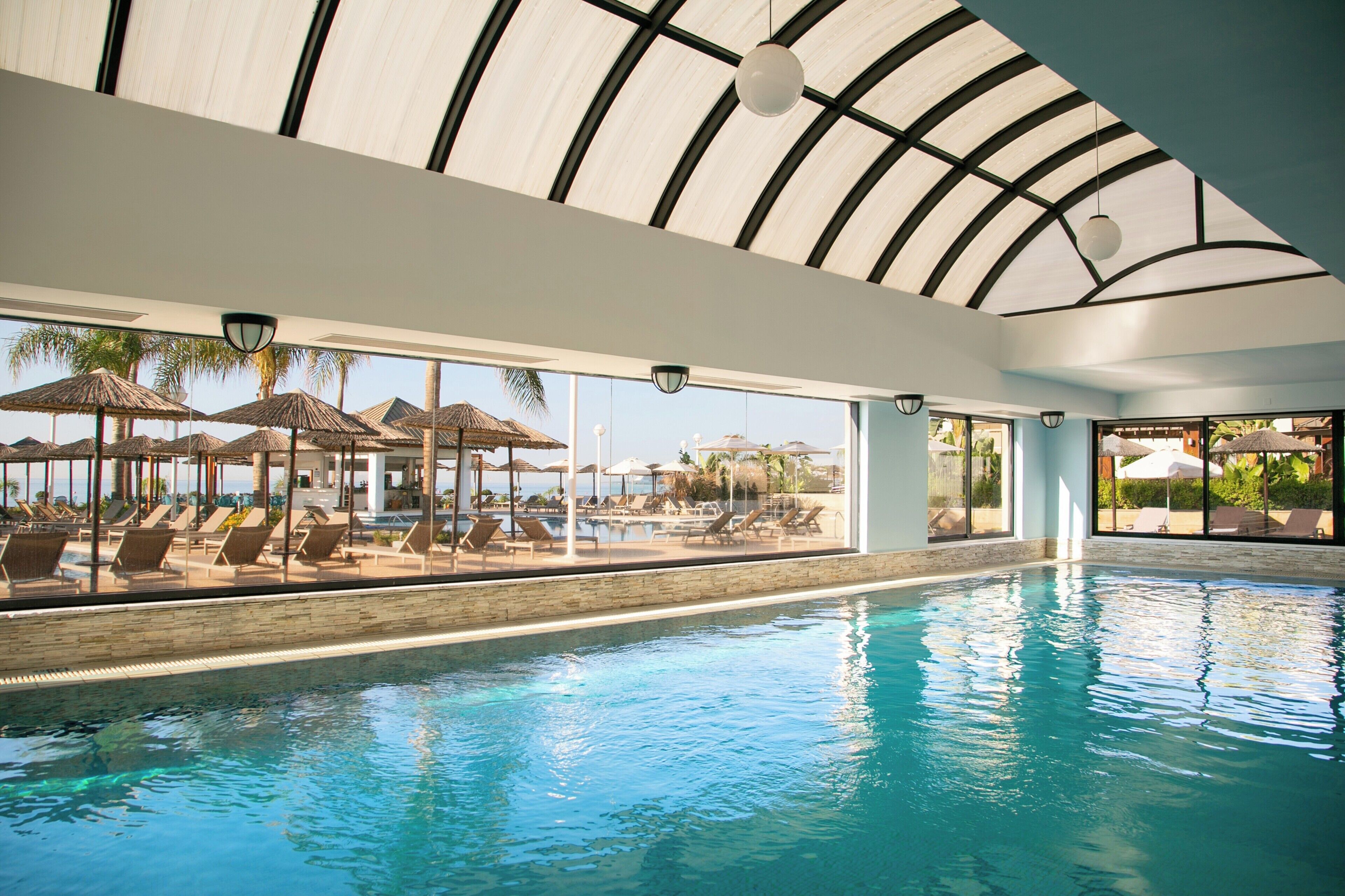 An indoor swimming pool with clear blue water, surrounded by lounge chairs and umbrellas, under a large glass ceiling.