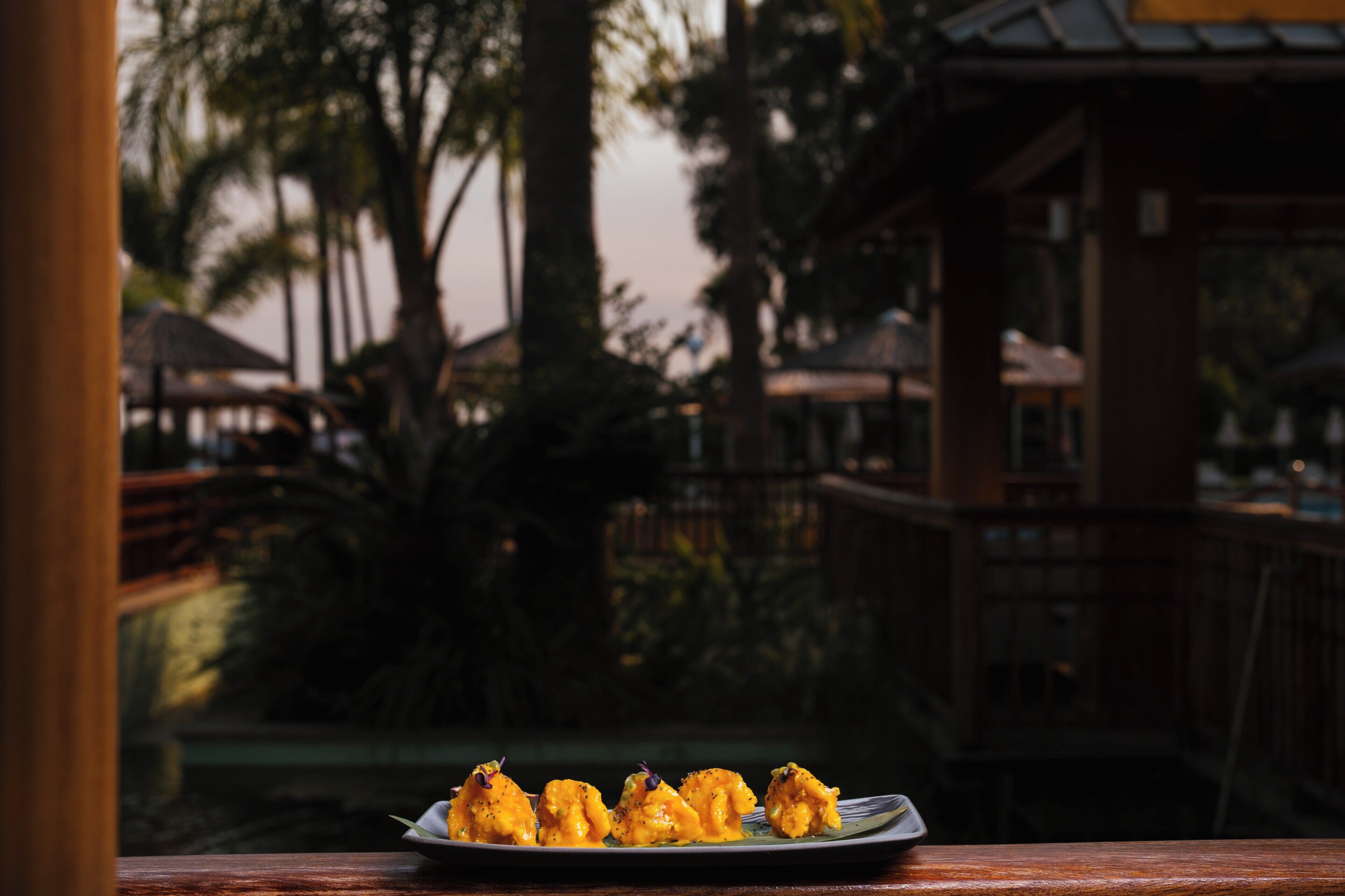 A wooden table with a plate of food, set against a blurred background of a building and trees.