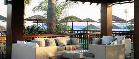 A covered outdoor seating area with wicker furniture and umbrellas, overlooking a beach.