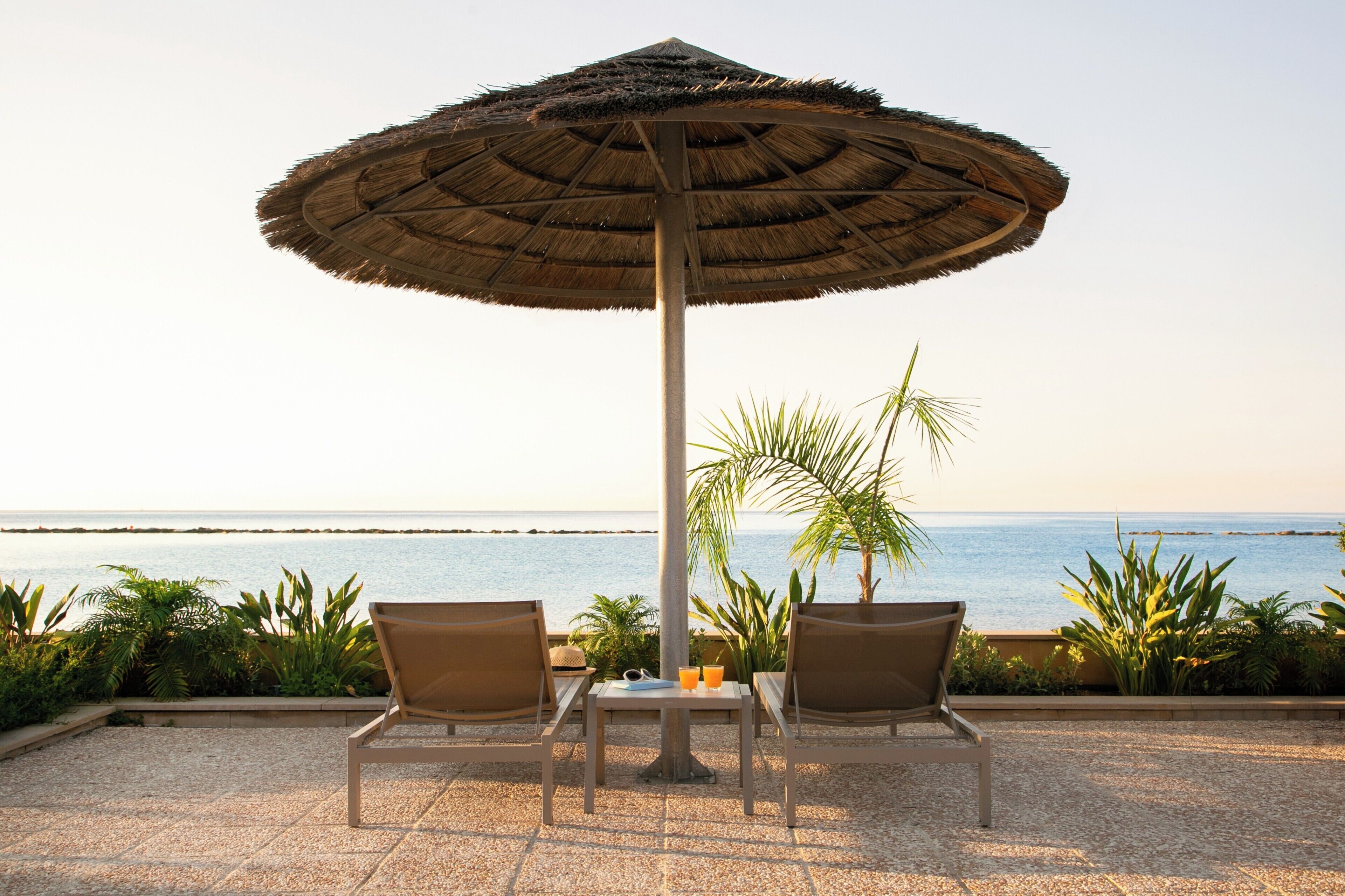 Deux fauteuils de jardin placés sous un grand parasol, face à une mer calme dont l’horizon est dégagé.
