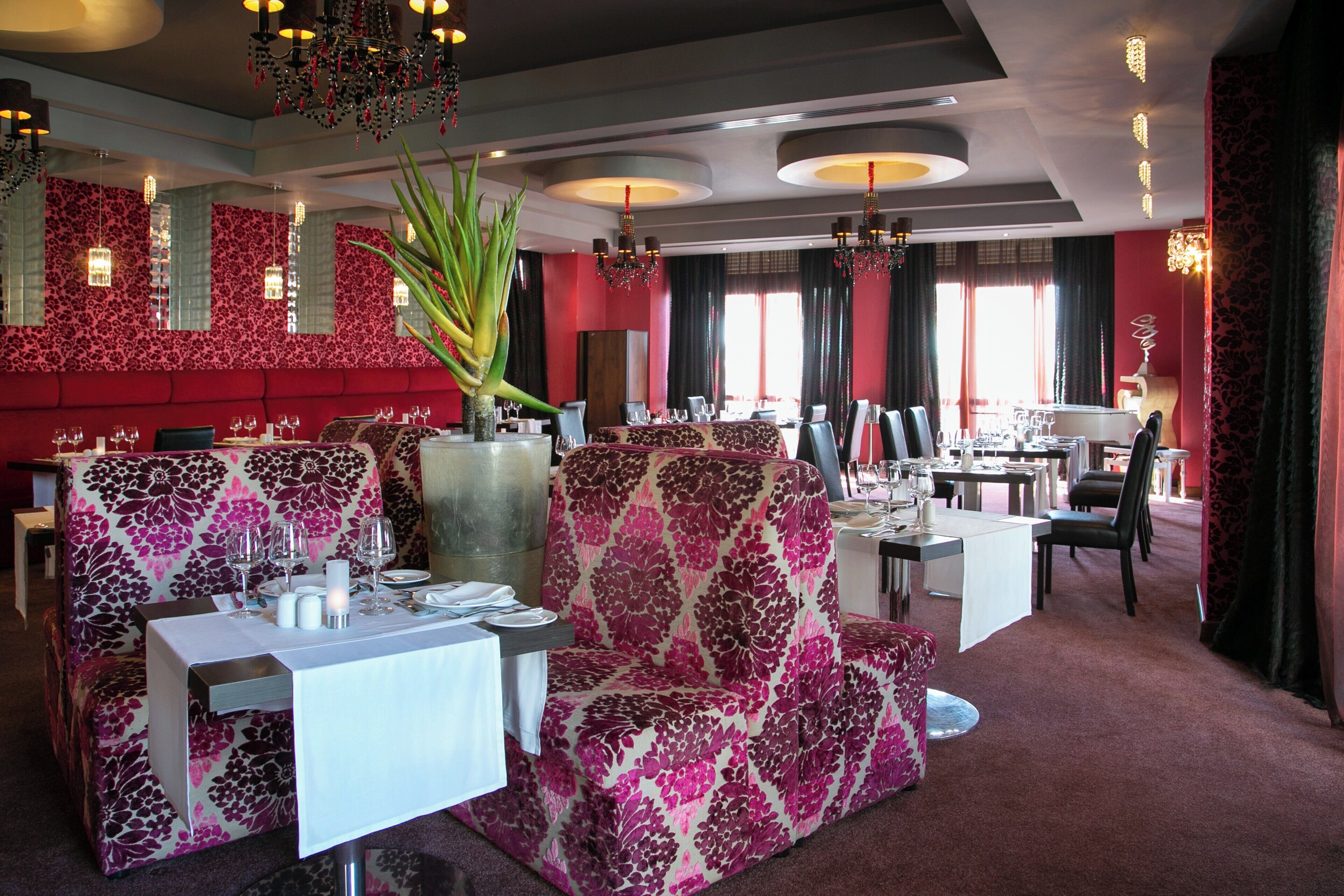 A restaurant interior with red walls, floral patterned furniture, and multiple dining tables set for guests.