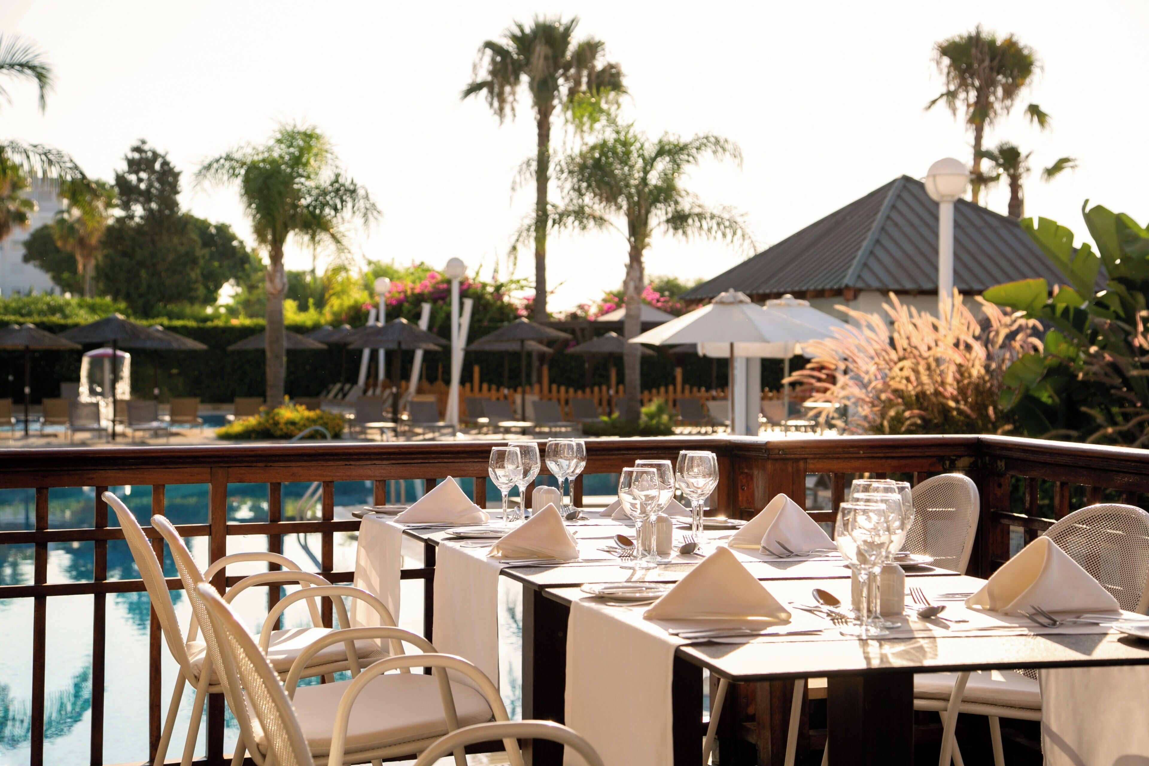 A dining area with tables set for two, overlooking a pool with palm trees and umbrellas in the background.