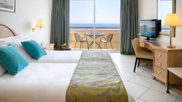 Quarto de hotel com cama, escrivaninha com TV, cadeira, mesa pequena e vista para o oceano através de amplas janelas.