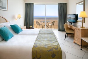 Quarto de hotel com cama, escrivaninha com TV, cadeira, mesa pequena e vista para o oceano através de amplas janelas.
