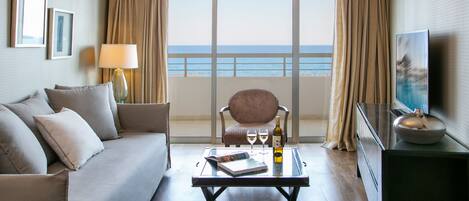 Quarto de hotel com varanda, um sofá, uma cadeira, uma mesa de centro com um livro e uma garrafa, e uma TV sobre um suporte.