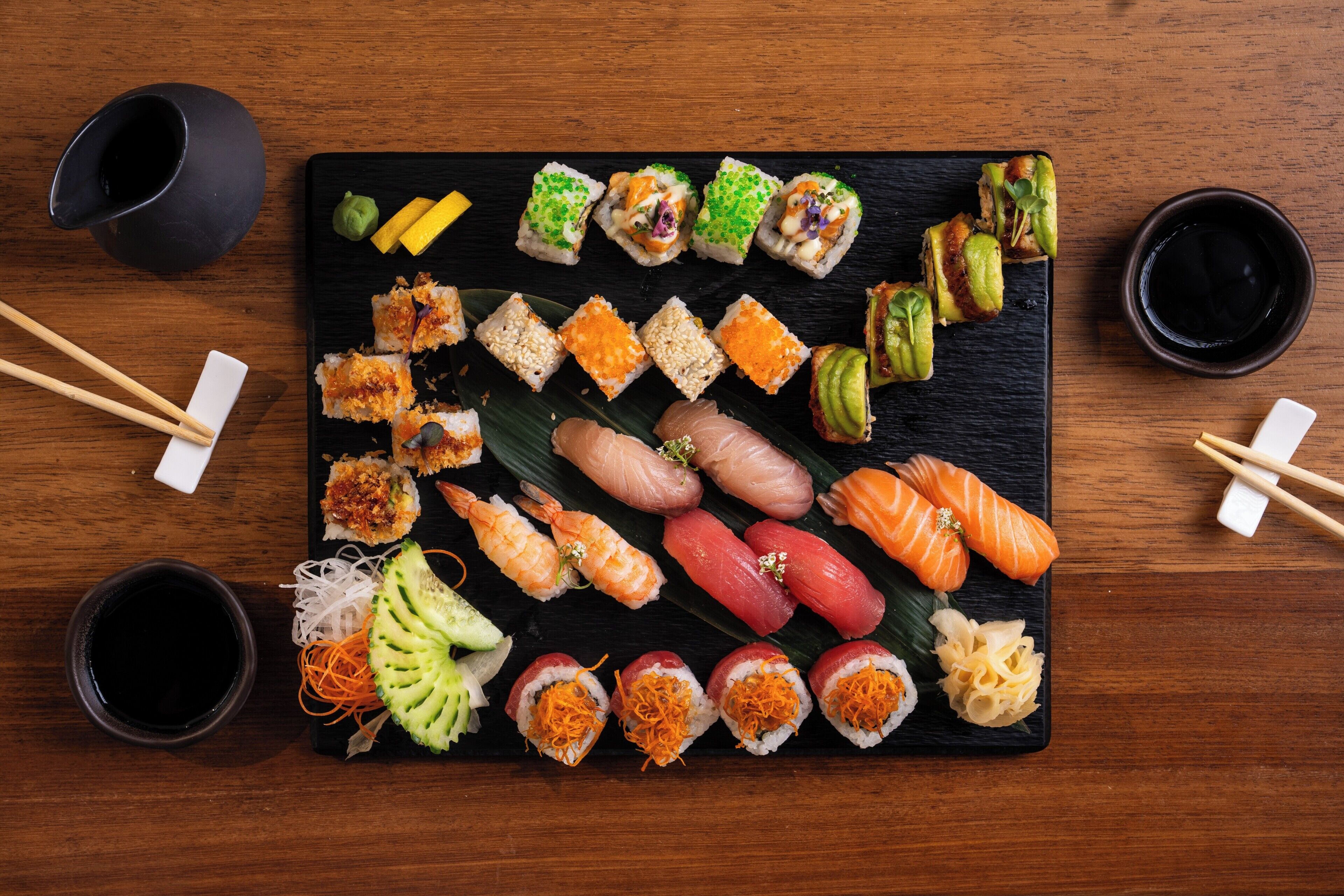 A variety of sushi on a black slate plate, with chopsticks and two black cups on a wooden table.