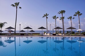 A poolside area with lounge chairs and umbrellas, a clear view of the sea, and a tiled pool reflecting the surroundings. - Atlantica Miramare Beach (Limassol)