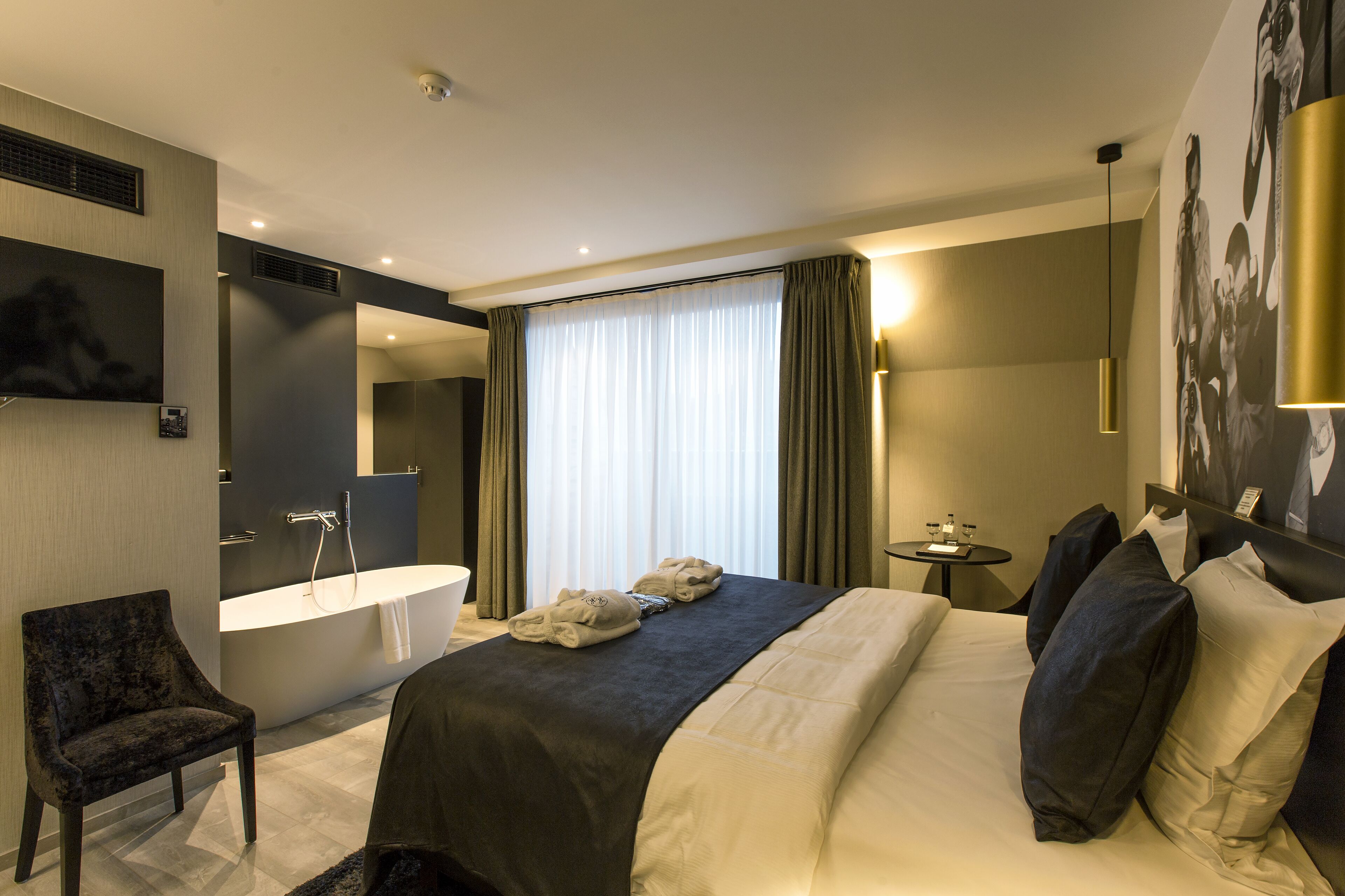 A modern hotel room with a large bed, a sitting area with a chair, a bathtub, and a small table with amenities.