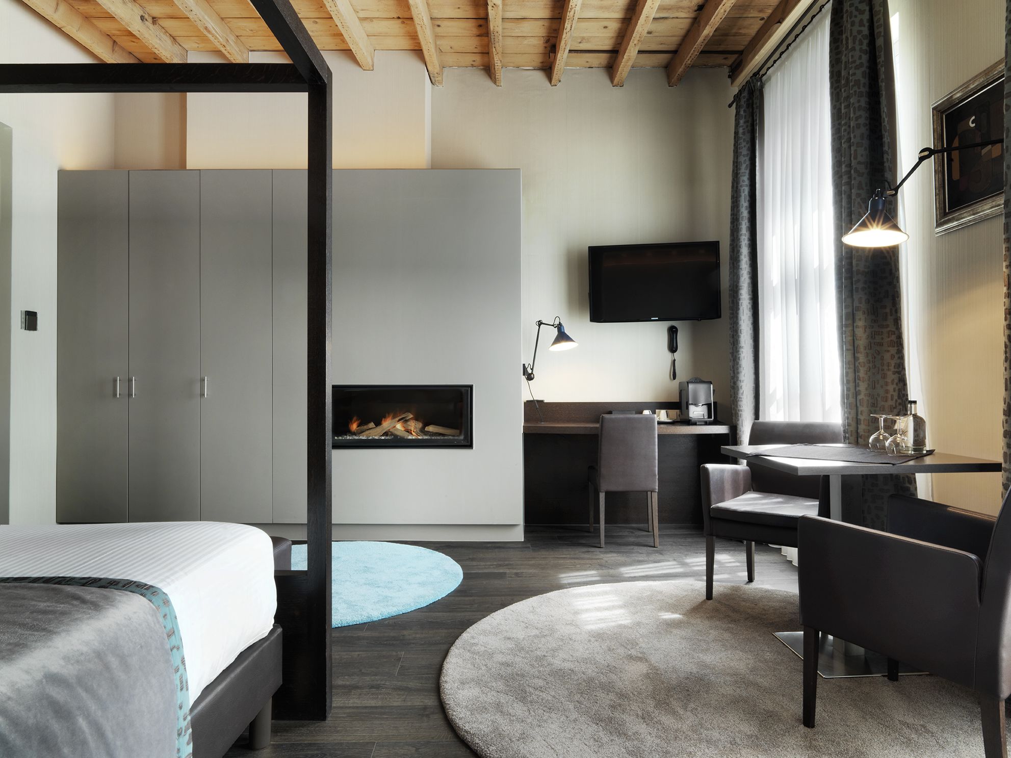 Quarto de hotel moderno com cama, escrivaninha, poltrona, televisão e lareira.