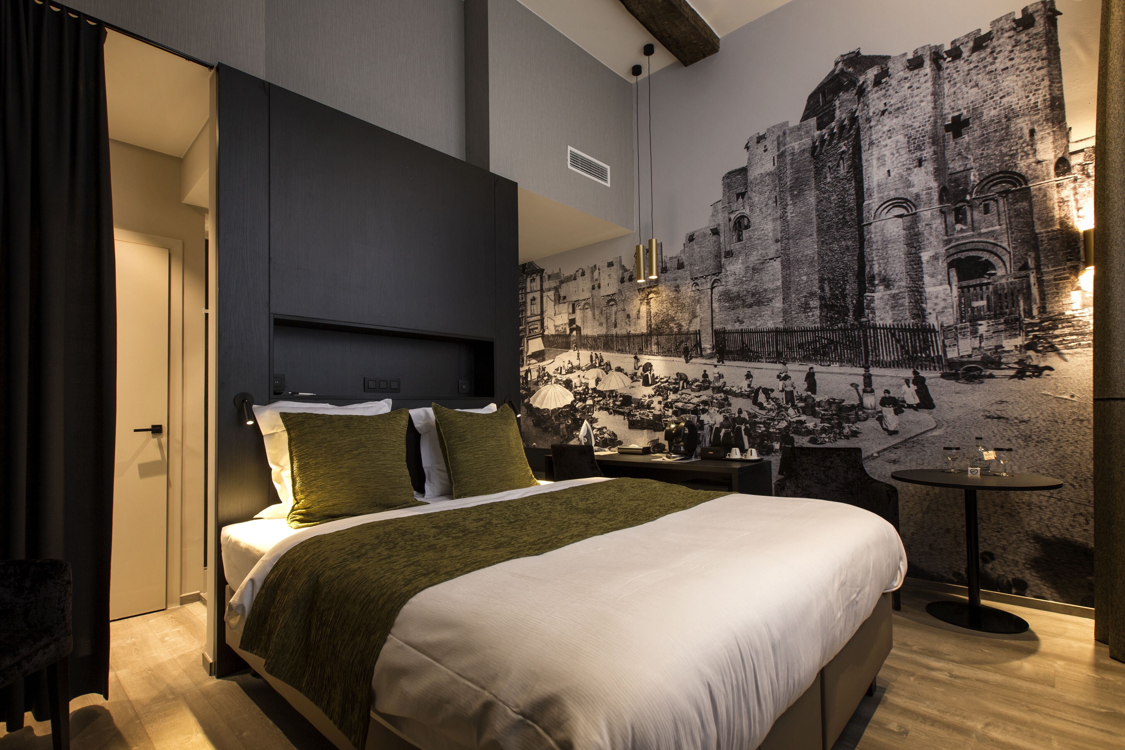 Une chambre d’hôtel moderne avec un grand lit, une petite table et une fresque représentant un bâtiment historique sur le mur.