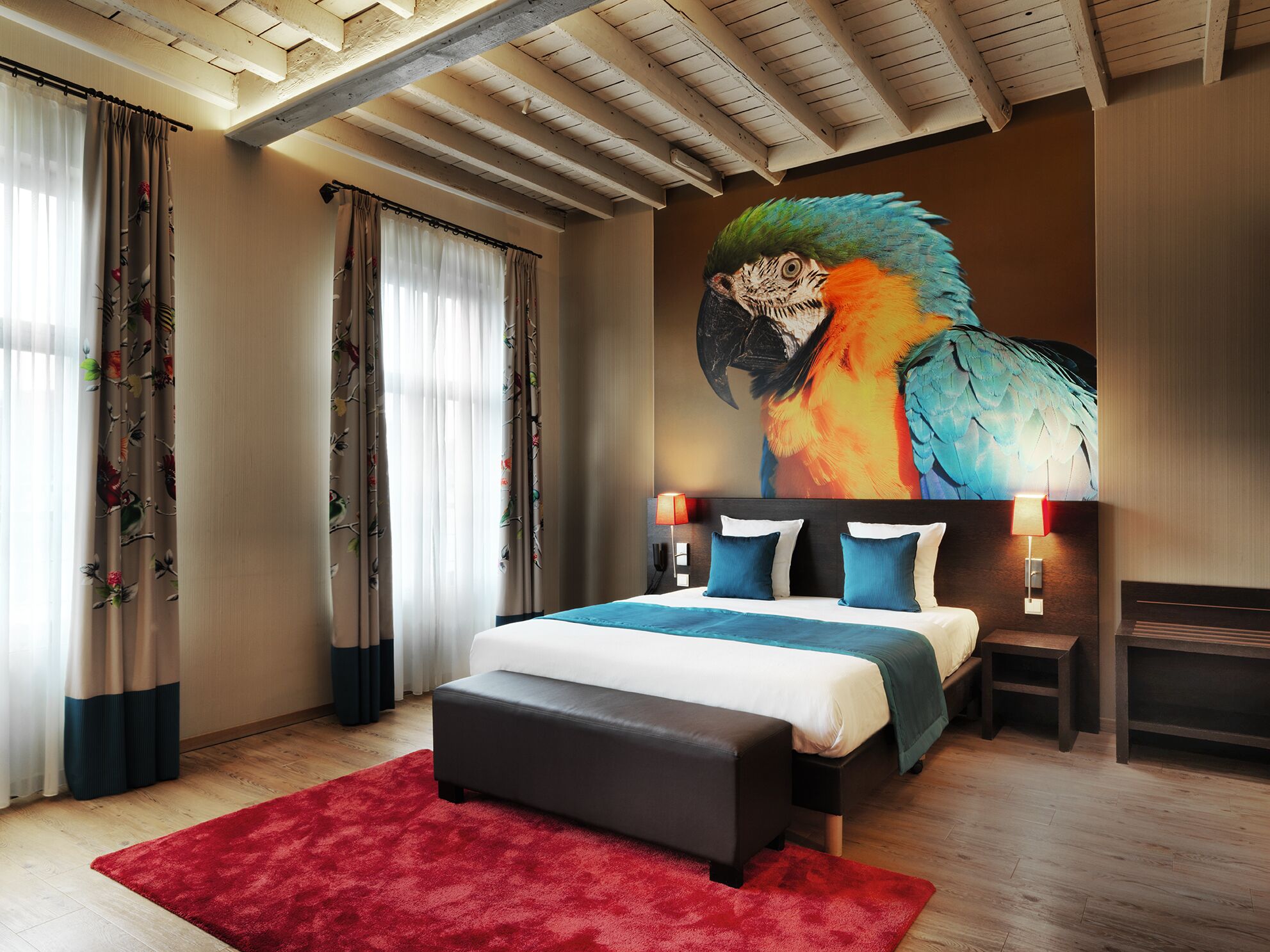 A modern bedroom with a large bed, a red rug, wooden flooring, and a mural of a parrot.