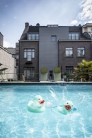 Una piscina con patitos hinchables, sillas de descanso y un edificio con ventanas y un balcón al fondo.