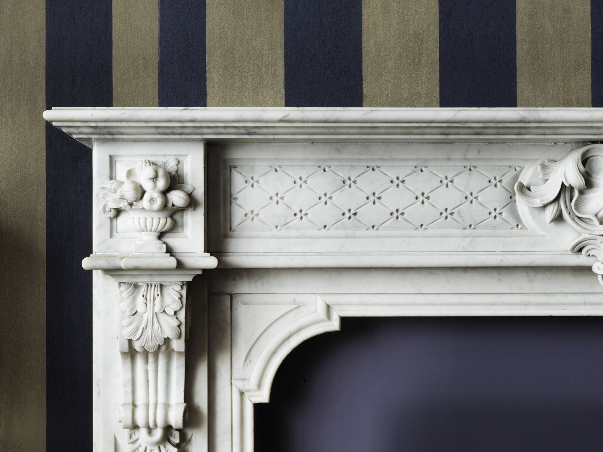 A white marble fireplace with intricate carvings and a decorative cornice against a striped wall.
