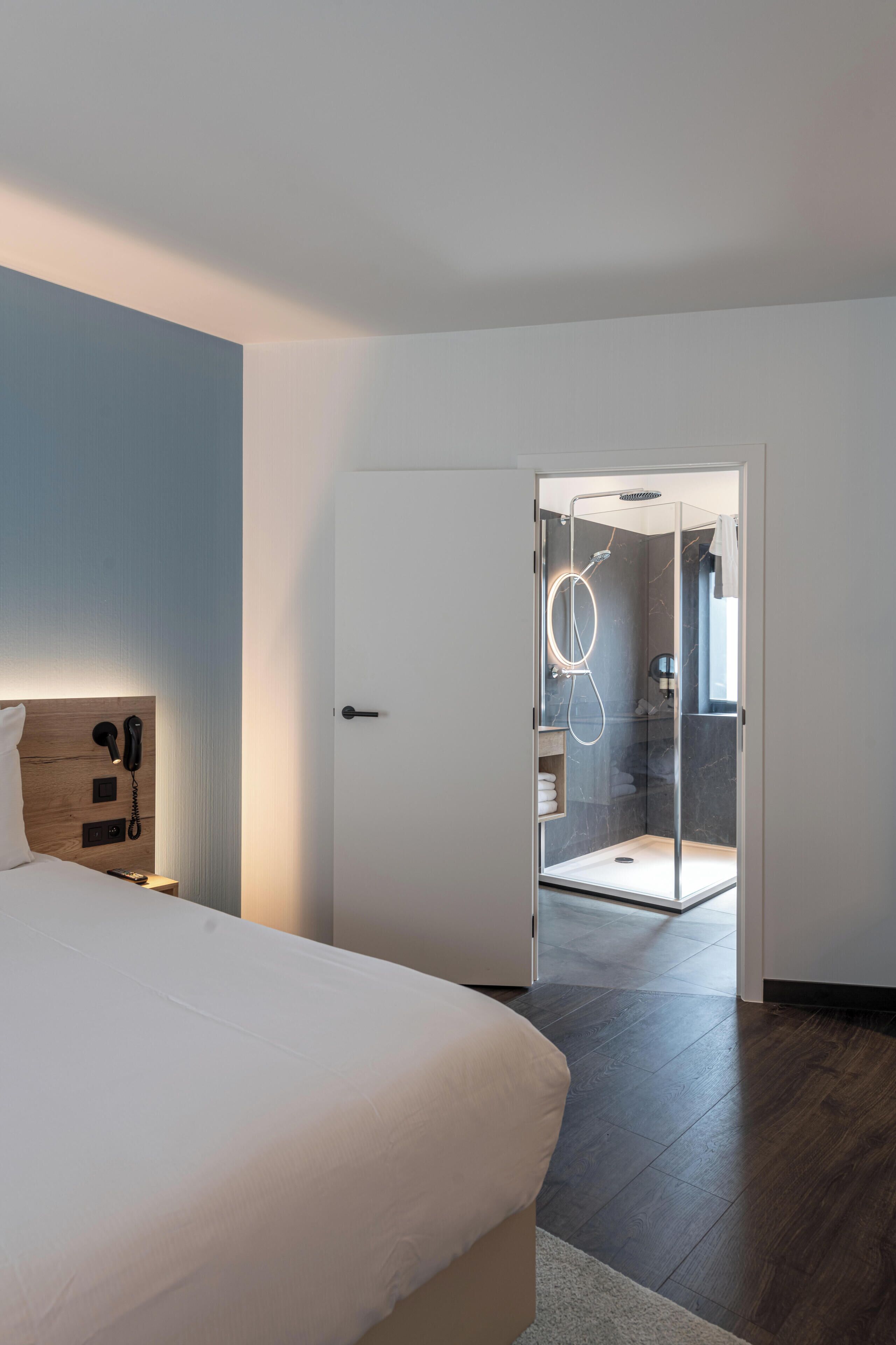 Une chambre d’hôtel moderne dotée d’un grand lit, d’une tête de lit en bois, d’un téléphone sur la table de chevet et d’une cabine de douche en verre.