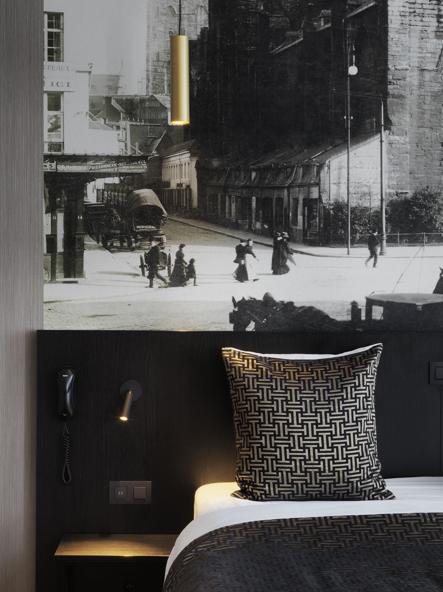 A hotel room with a large poster of a street scene, a patterned pillow, a bedside table, and a hanging light fixture.