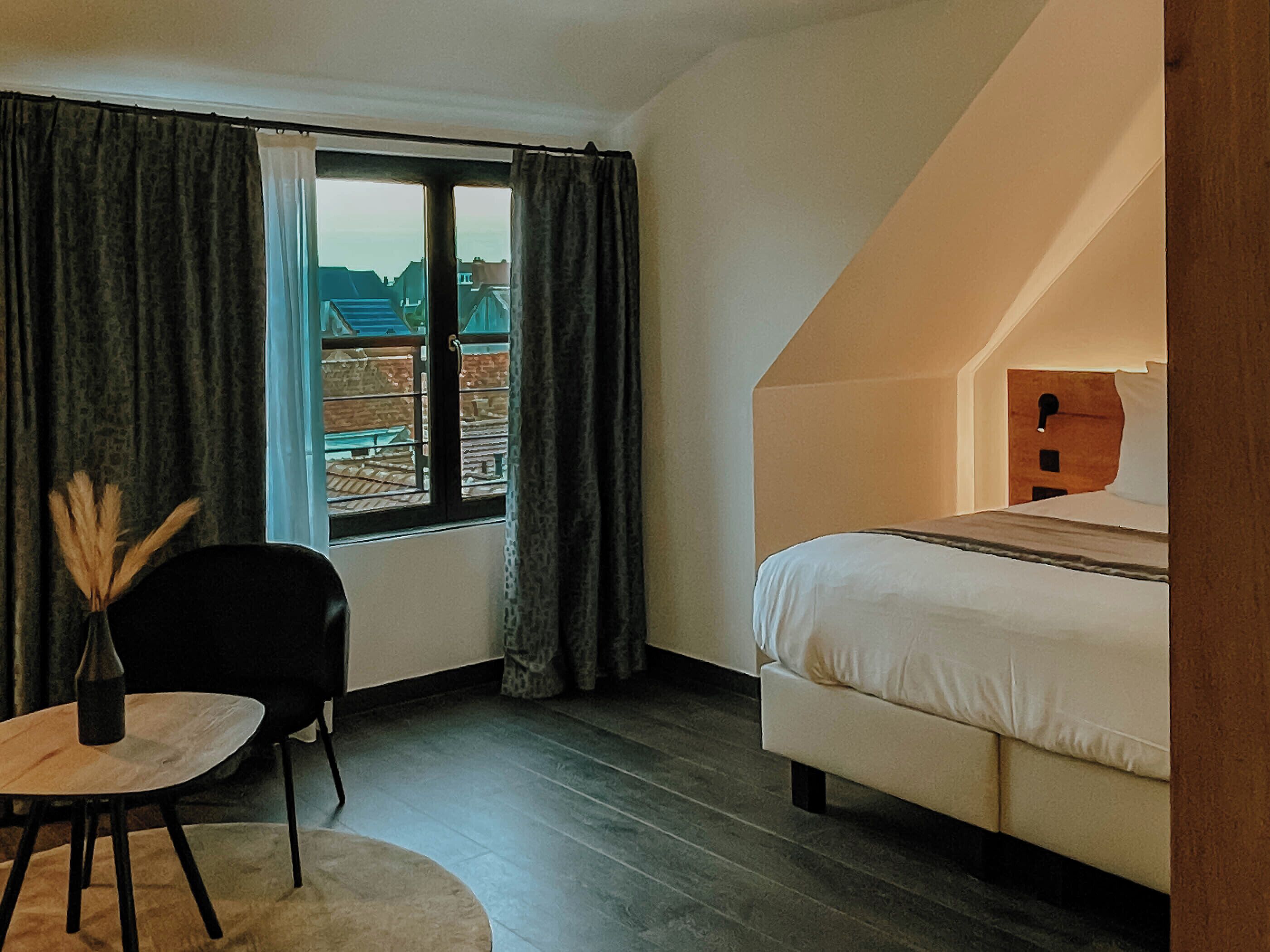 Une chambre à coucher avec un lit, une chaise, une petite table et une fenêtre avec des rideaux.