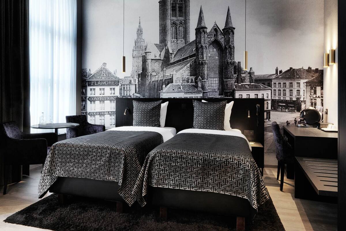 Habitación de hotel con dos camas, un ventanal con cortinas, un mural de paisaje urbano en la pared y un escritorio con una silla.