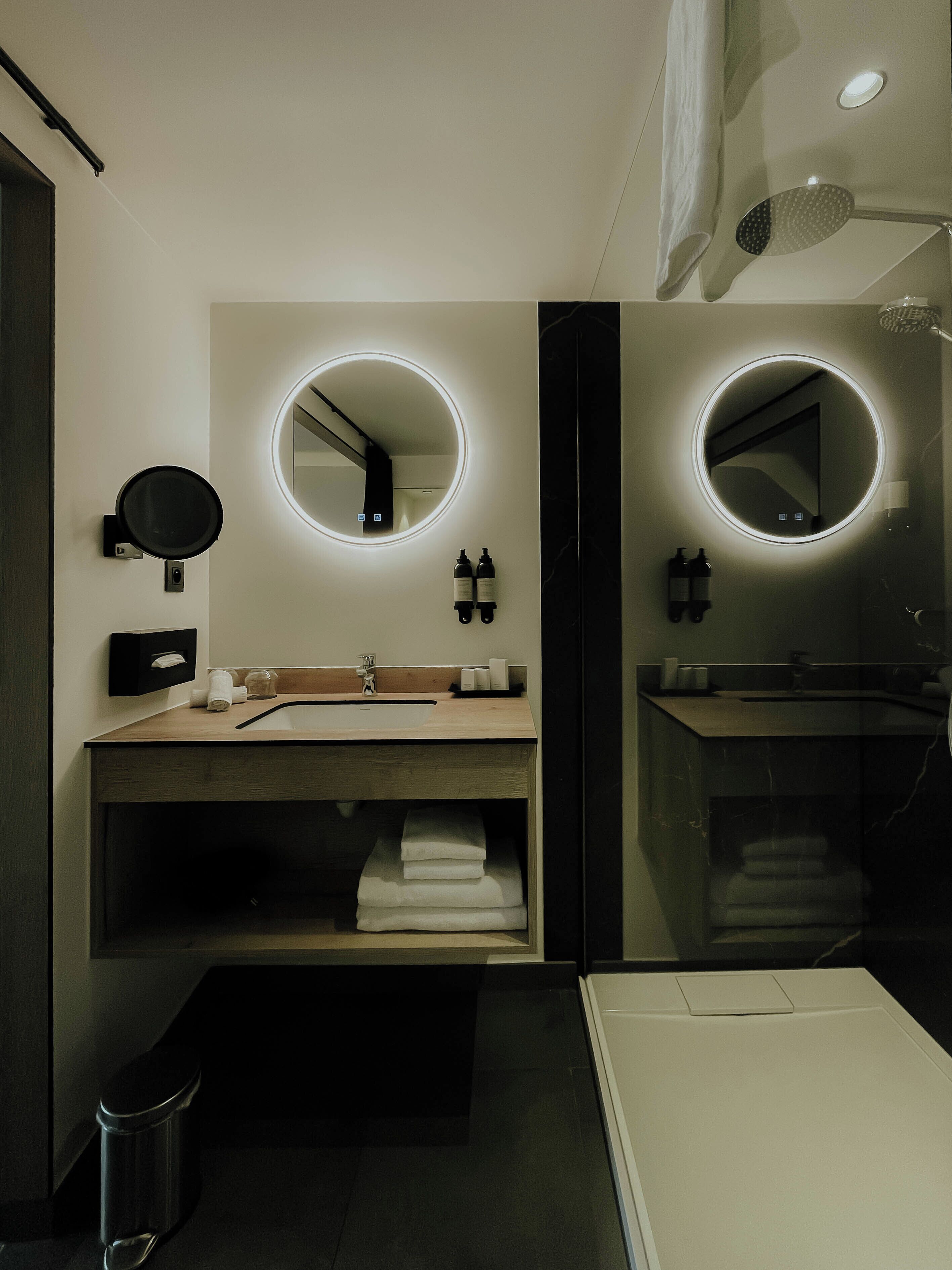 Un baño moderno con dos lavamanos, cada uno con un espejo redondo e iluminación debajo del mueble. Hay toallas y artículos de aseo en los lavamanos, y se ve un cubo de basura.