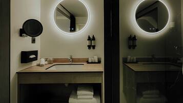 Un baño moderno con dos lavamanos, cada uno con un espejo redondo e iluminación debajo del mueble. Hay toallas y artículos de aseo en los lavamanos, y se ve un cubo de basura.