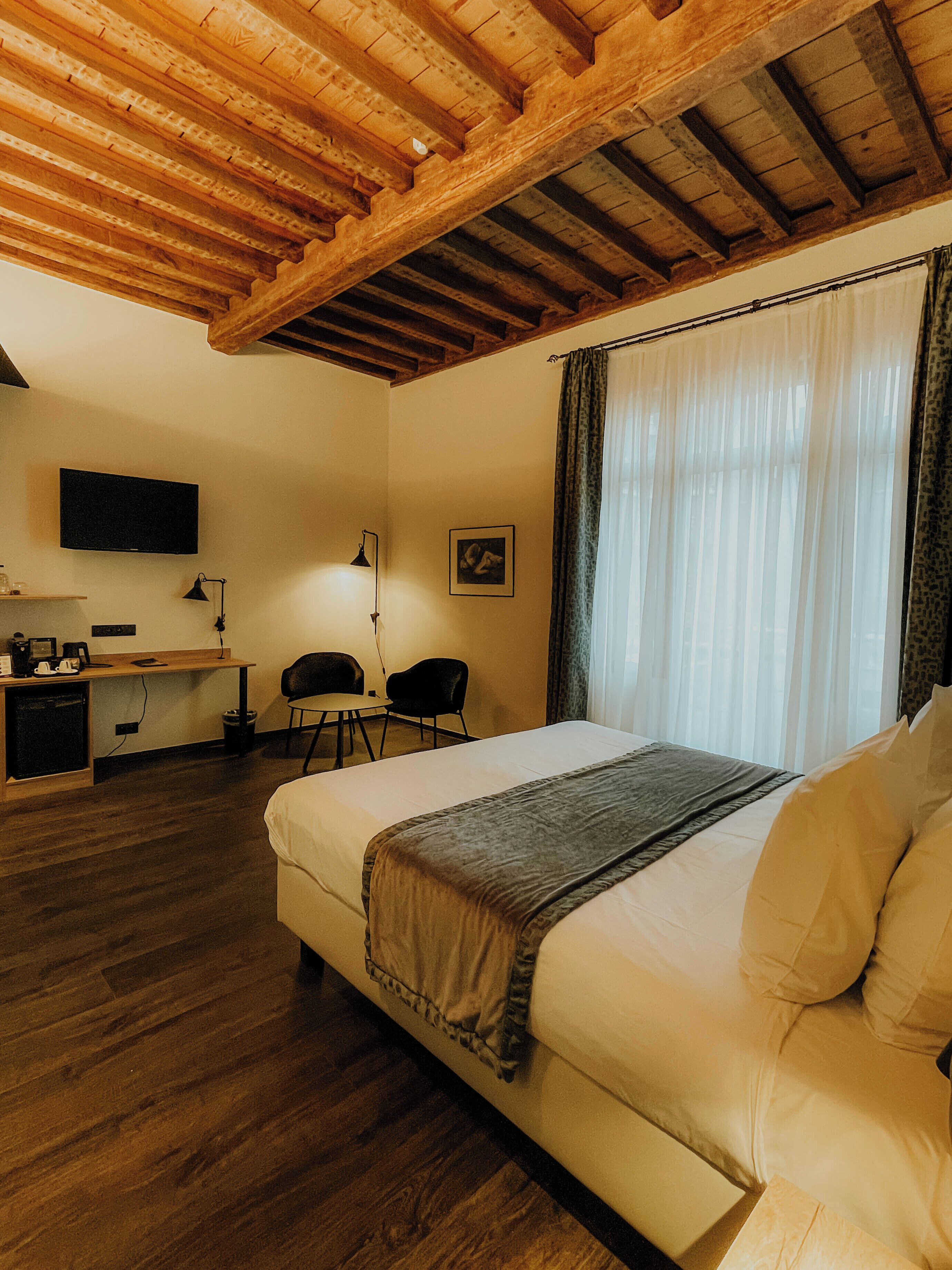 A hotel room with a wooden ceiling, a bed, a desk with a chair, a television, and a window with curtains.