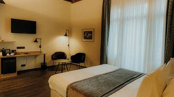 A hotel room with a wooden ceiling, a bed, a desk with a chair, a television, and a window with curtains.