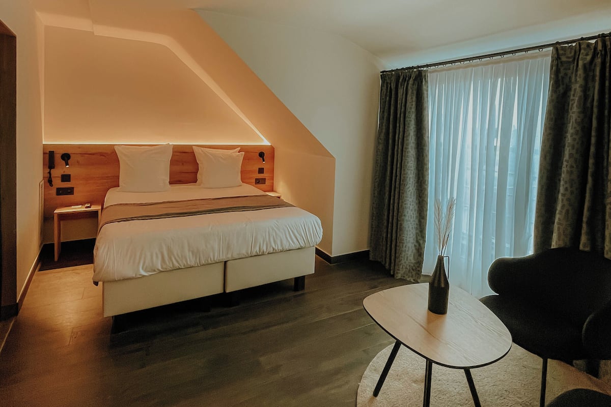Habitación de hotel con cama, mesitas de noche, una pequeña mesa, una silla y una ventana con cortinas.