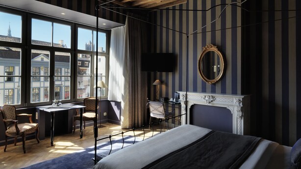 Habitación de hotel con una cama grande, un escritorio, una silla, chimenea, televisión y ventanas amplias con vistas a la ciudad.