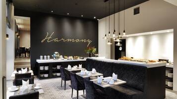A modern restaurant interior with a black wall featuring the word "Harmony" in gold lettering, a bar area with a variety of food items, and a seating area with tables set for dining.