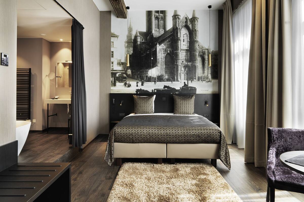 Una habitación de hotel moderna con una cama grande, baño y vistas a un edificio histórico.