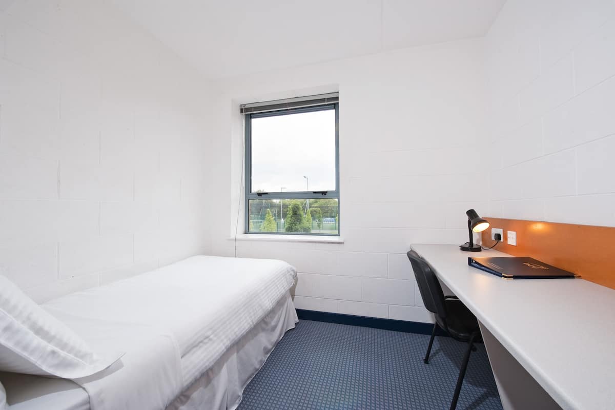 A small, clean room with a single bed, a desk with a chair, and a window.