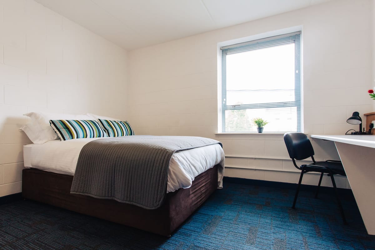 A bedroom with a bed, a desk, a chair, a window with blinds, and a small potted plant.
