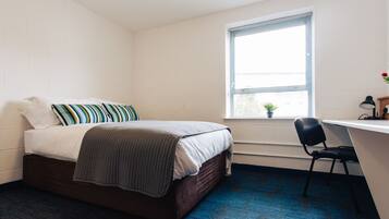 A bedroom with a bed, a desk, a chair, a window with blinds, and a small potted plant.