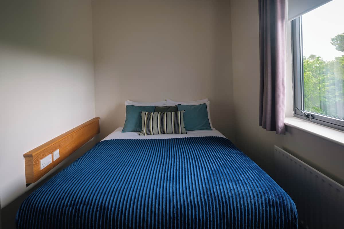 A neatly made bed with a blue striped bedspread and two pillows, a wooden headboard, and a window with curtains.
