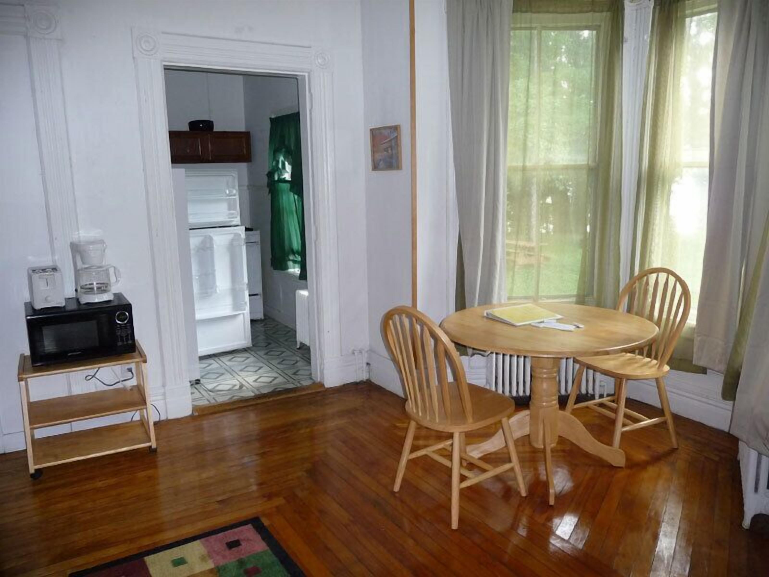 A dining area with a wooden table and chairs, a small shelf with appliances, and a view through a window with curtains.