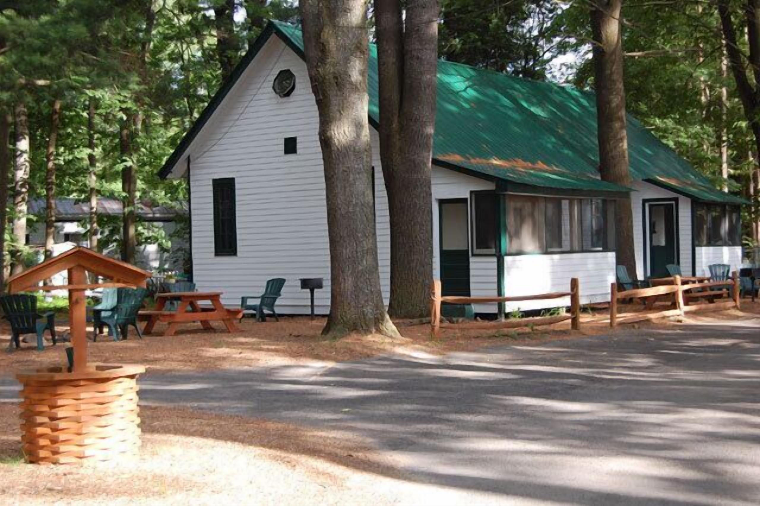 A white cabin with a green roof, surrounded by trees, and featuring outdoor seating and a picnic table.