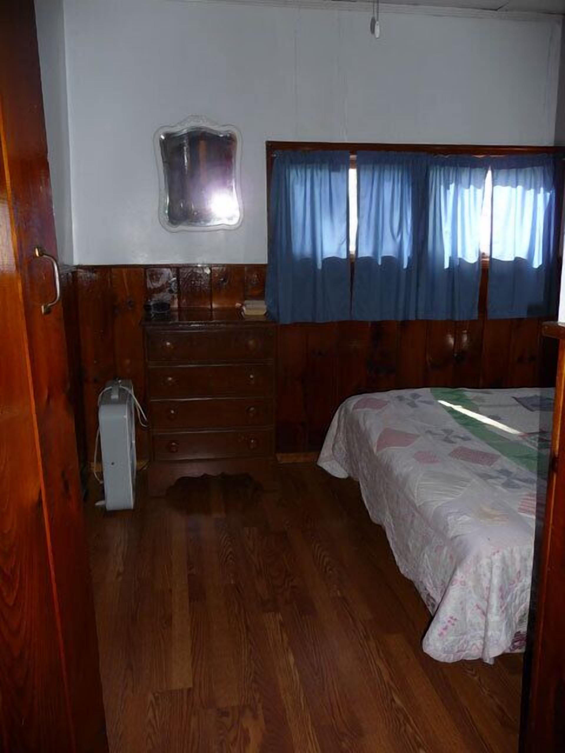 A bedroom with a wooden dresser, a bed with a patterned bedspread, a window with blue curtains, and a small mirror above the dresser.