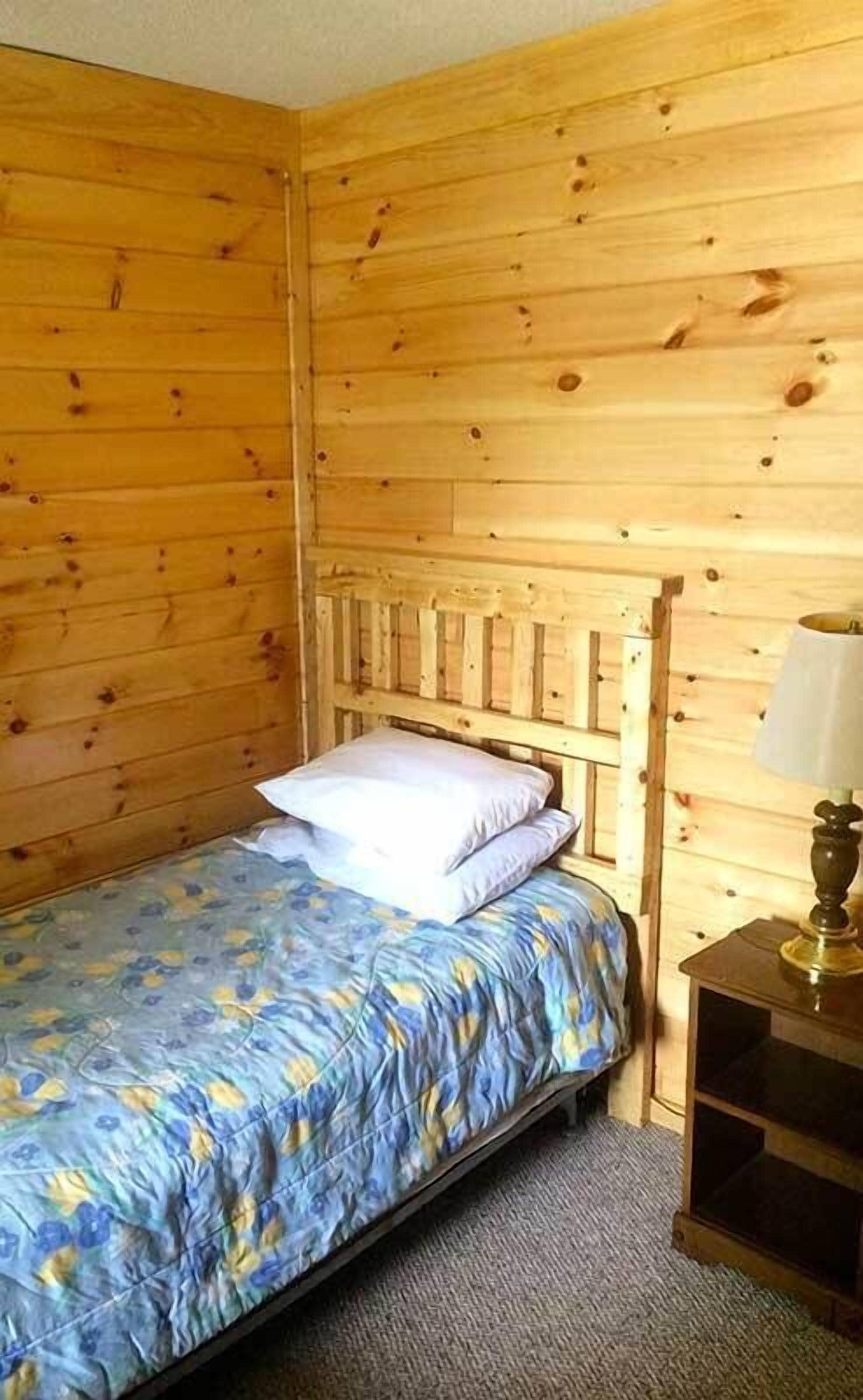 A single bed with a floral bedspread in a room with wooden walls and a nightstand with a lamp.