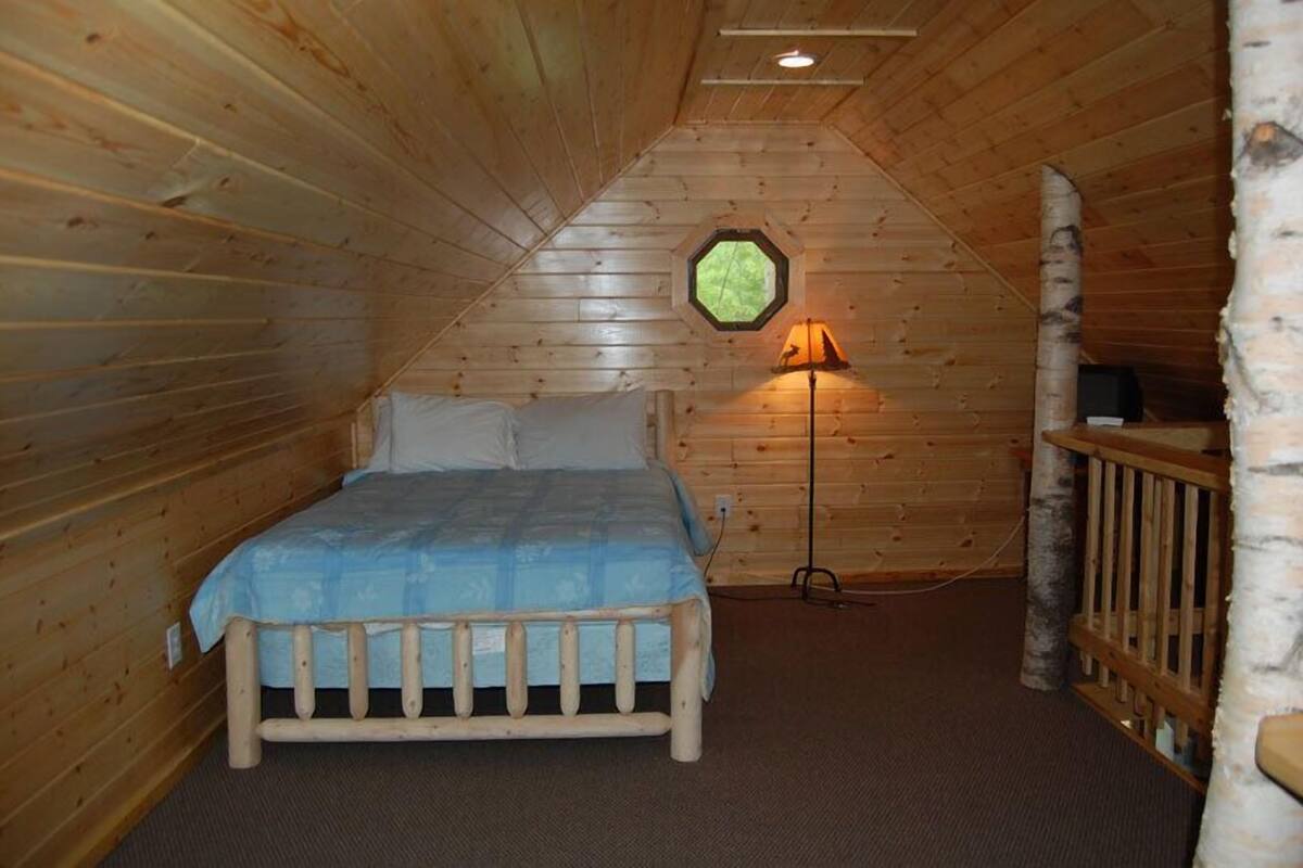 A wooden cabin bedroom with a bed, a lamp, and a window.