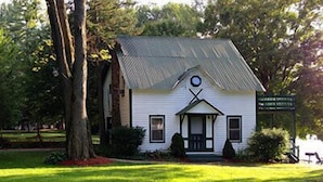 Un petit bâtiment blanc, doté d’un toit à pignon et d’un porche, entouré d’arbres et d’herbe.