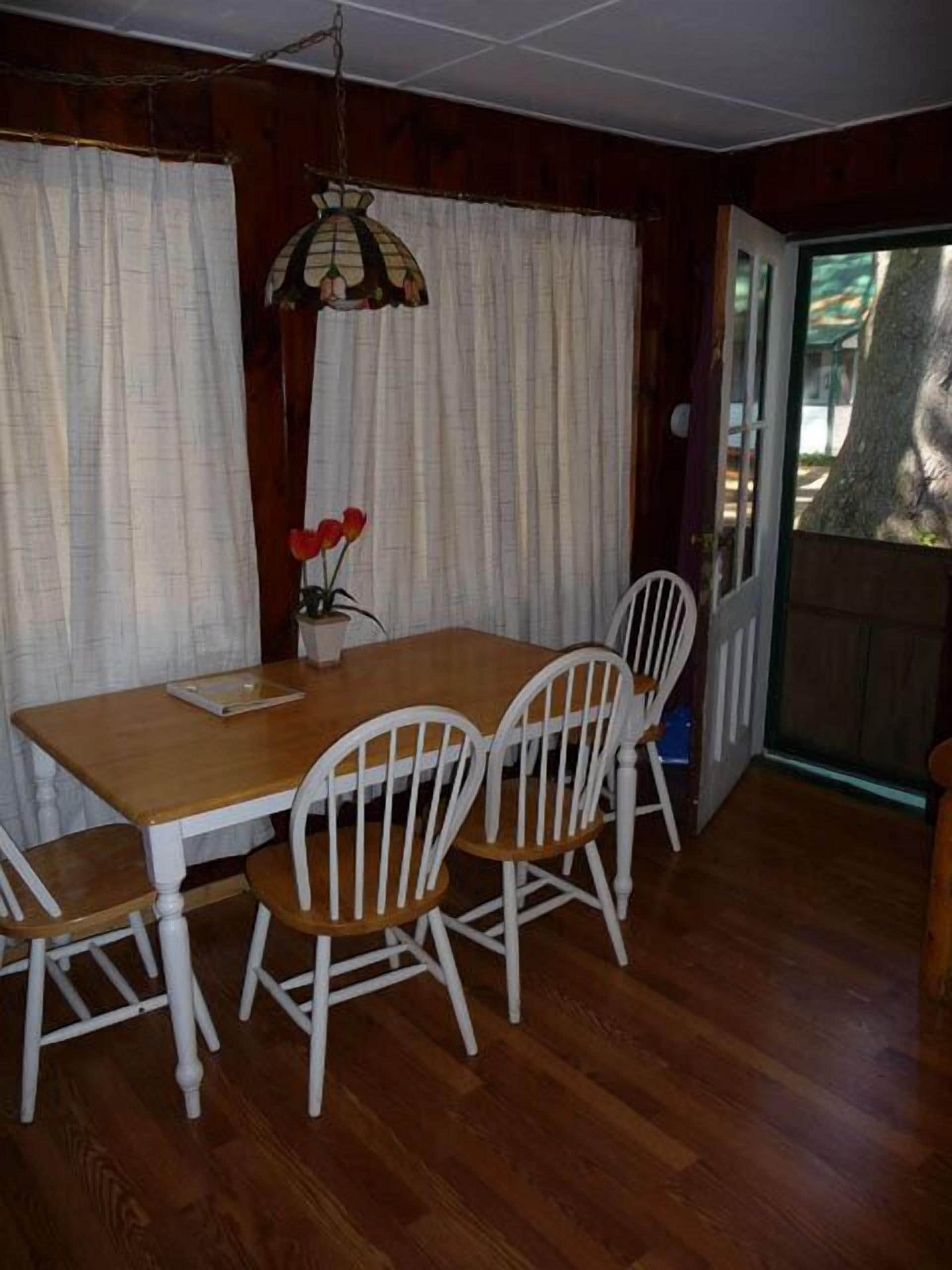 A dining area with a wooden table and four white chairs, a hanging lamp, and a door leading outside.