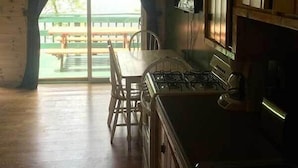 L’intérieur d’un chalet en bois comprend une salle à manger avec une table et des chaises, une cuisine équipée de placards et une vue sur un espace extérieur verdoyant, accessible par une porte coulissante.
